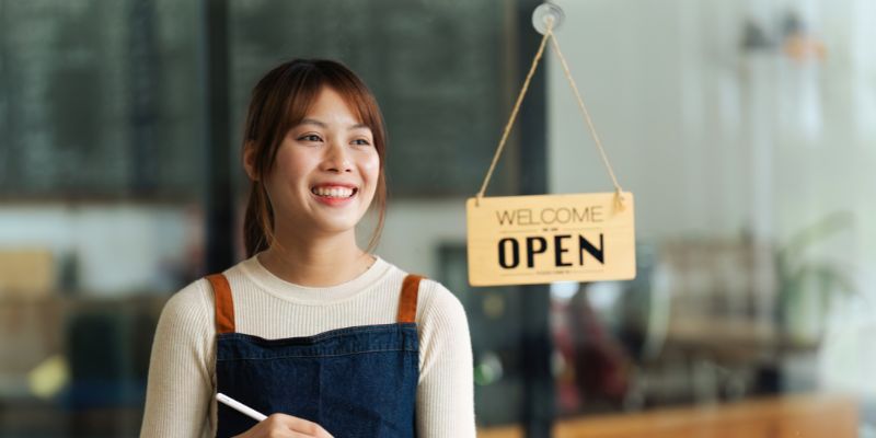 Seorang pelanyang F&B perempuan sedang berdiri di hadapan kafe sambil tersenyum.